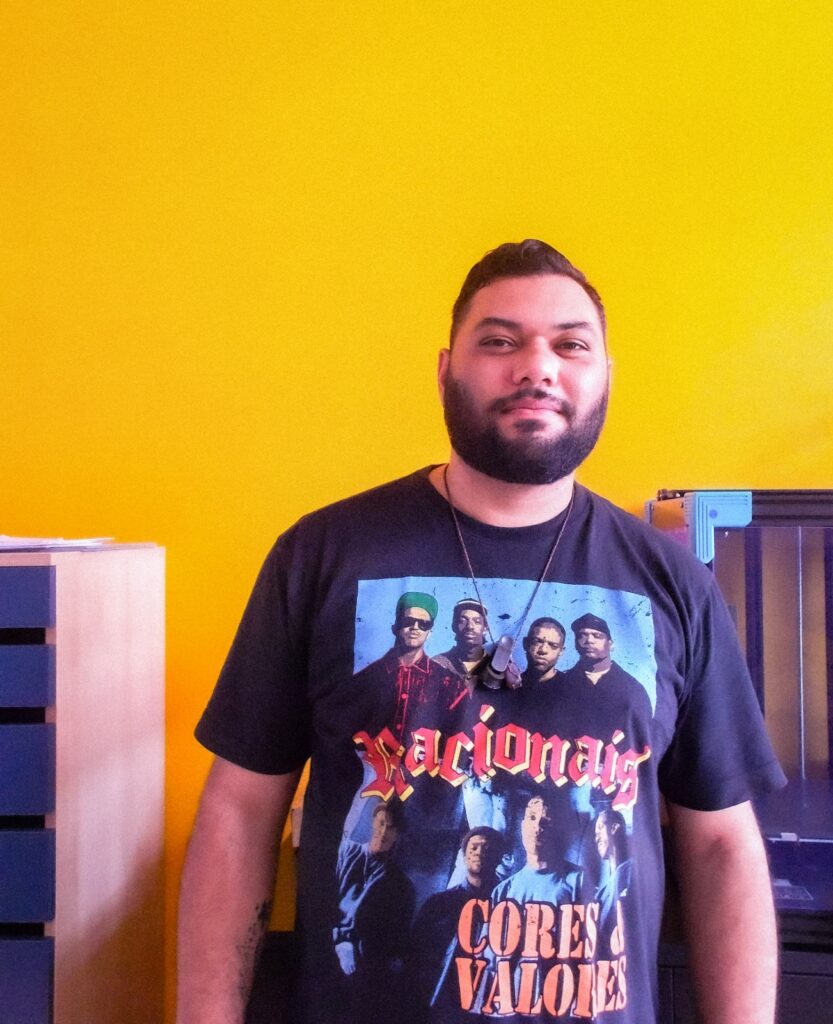 Rafael Braga ostentando sua camiseta do Racionais em um estúdio com parede amarela